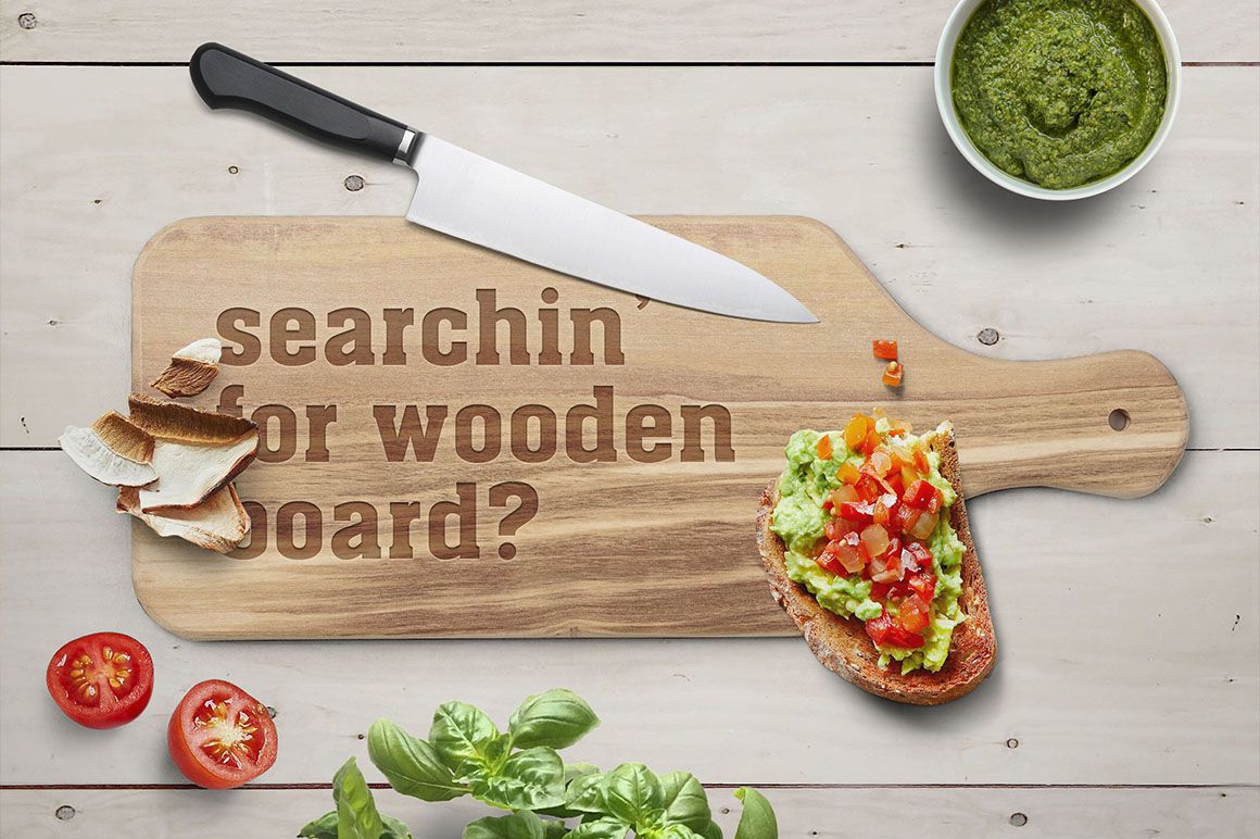 Top view of a rectangular wooden board mockup with engraved text, knife, bread, tomatoes, and bowl of green pesto.