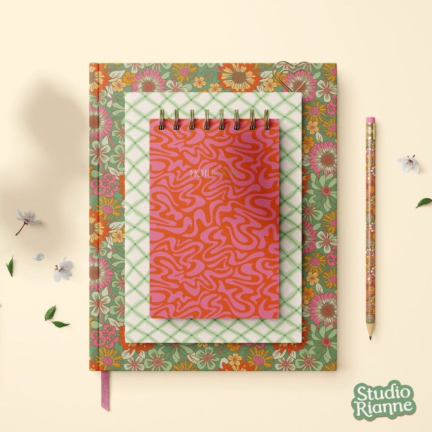 Stationery set mockup featuring a spiral notebook with a red abstract cover stacked on patterned books, with a floral pencil nearby.