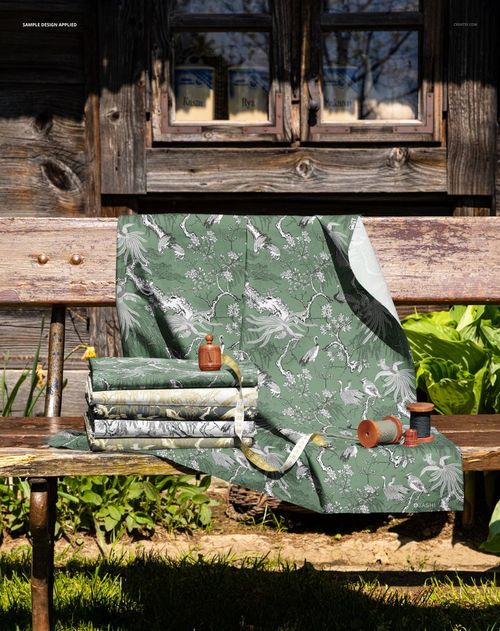 PSD mockup showing folded and draped fabrics with nature-inspired designs on a wooden bench in a garden setting.