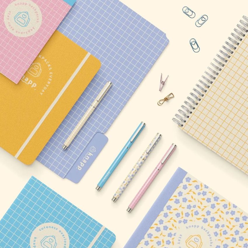 Stationery set mockup featuring grid-patterned notebooks, pastel pens, and paper clips arranged on a light surface.