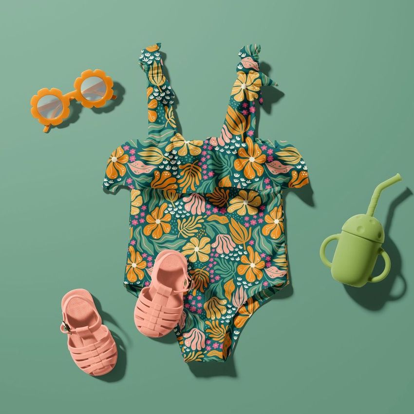Baby swimsuit mockup with a colorful tropical floral pattern, styled with pink sandals, orange sunglasses, and a green sippy cup on a green background.