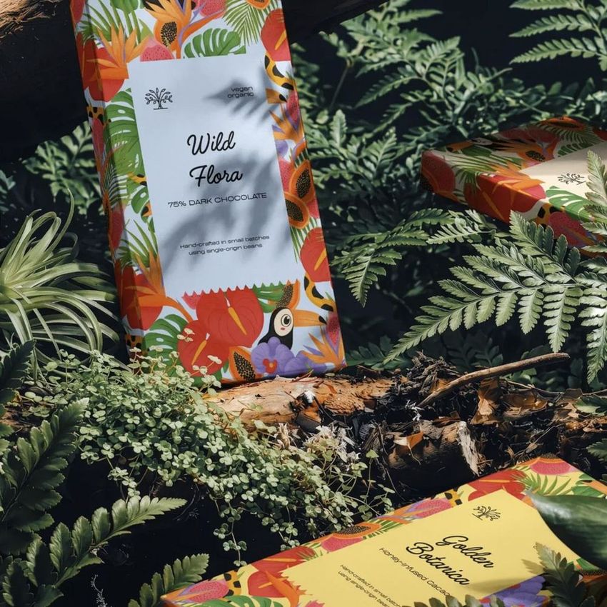 Chocolate box mockup featuring colorful botanical packaging designs, displayed among green ferns and moss in a forest setting.