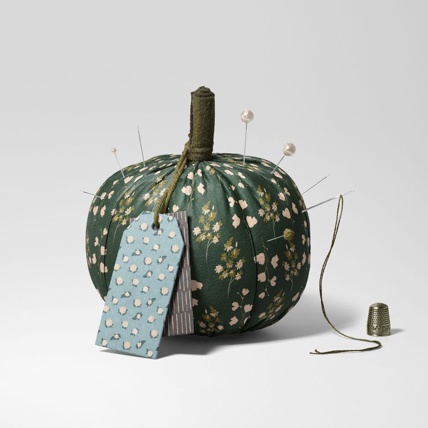Fabric pumpkin mockup with a green floral pattern, sewing pins inserted on top, and a blue patterned tag attached, set on a plain surface.