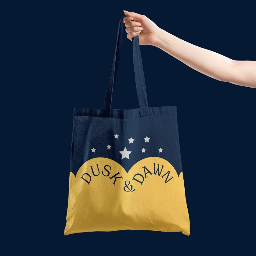Tote bag mockup featuring a hand holding a bag with a navy and yellow design, star graphics, and curved text overlay.