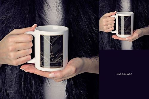 White ceramic mug with a geometric black and gold design held by a person wearing a dark fur garment, front view.