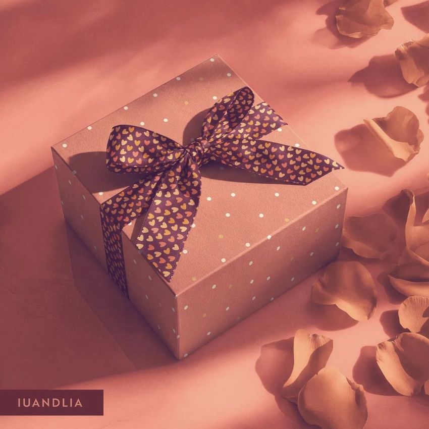 Square gift box mockup with a polka dot pattern and heart-patterned ribbon, surrounded by scattered rose petals on a pink surface.