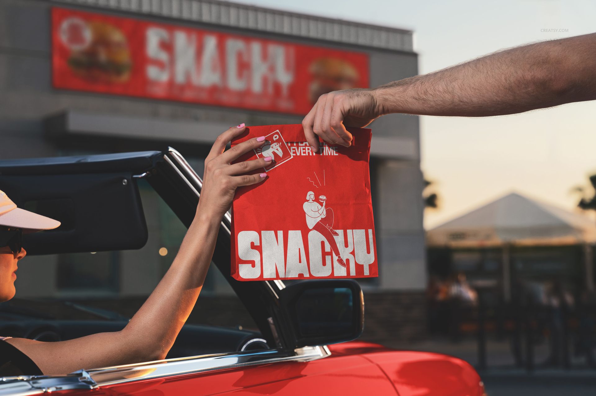 Person in a convertible receives a red fast food paper bag with bold white branding in a drive-through setting at sunset.