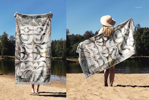 Mockup of a fringed beach towel featuring bold white text and leaf motifs, displayed by a lake on sandy ground.