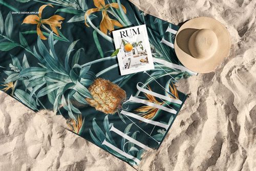 PSD mockup showing a rectangular beach towel with botanical print, placed on sandy beach with accessories beside.