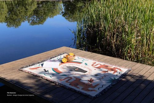 Fringed woven throw blanket mockup featuring illustrated tigers and leaves, laid out on a lakeside wooden platform.