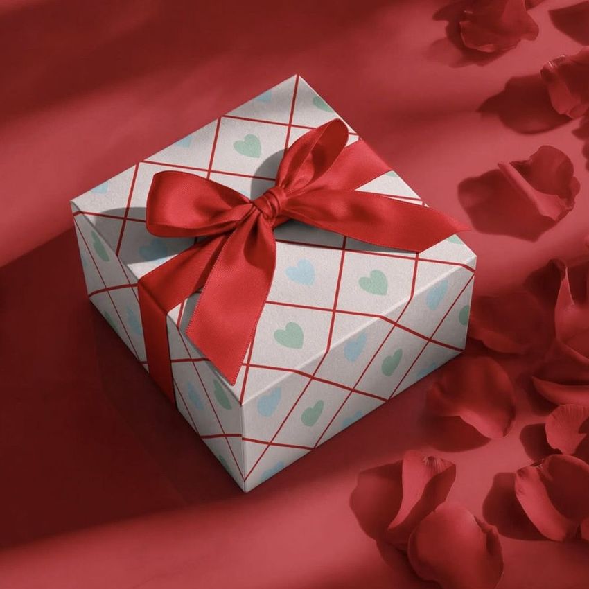 Square gift box mockup with a red ribbon, heart pattern, and grid lines, surrounded by scattered rose petals on a red surface.