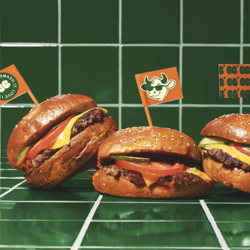 Burger mockup featuring three beef burgers with cheese, tomato, and lettuce, each topped with a branded flag on a green tiled surface.