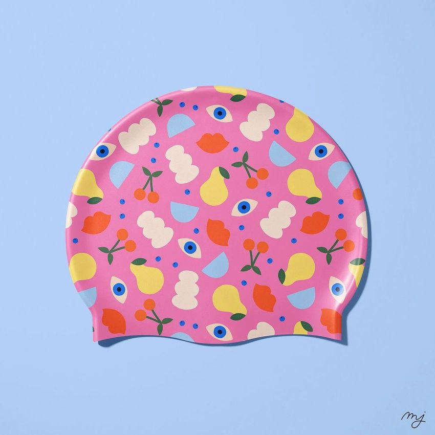 Swim cap mockup featuring a pink cap decorated with colorful abstract fruit and eye patterns on a light blue background.