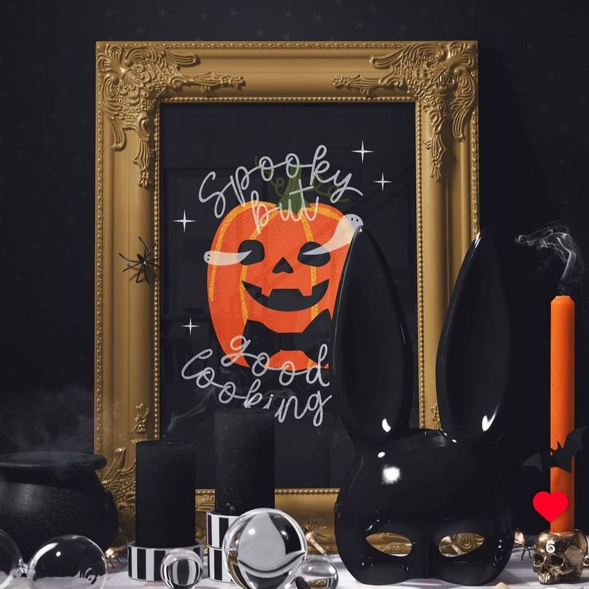 Halloween picture frame mockup featuring an ornate gold frame with a pumpkin art print, surrounded by black candles, an orange candle, and a black rabbit mask on a dark patterned background.
