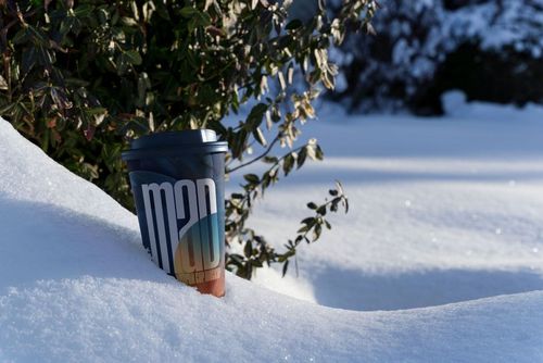 Winter scene featuring a paper cup mockup with a blue lid and multicolored pattern placed in snowy landscape.