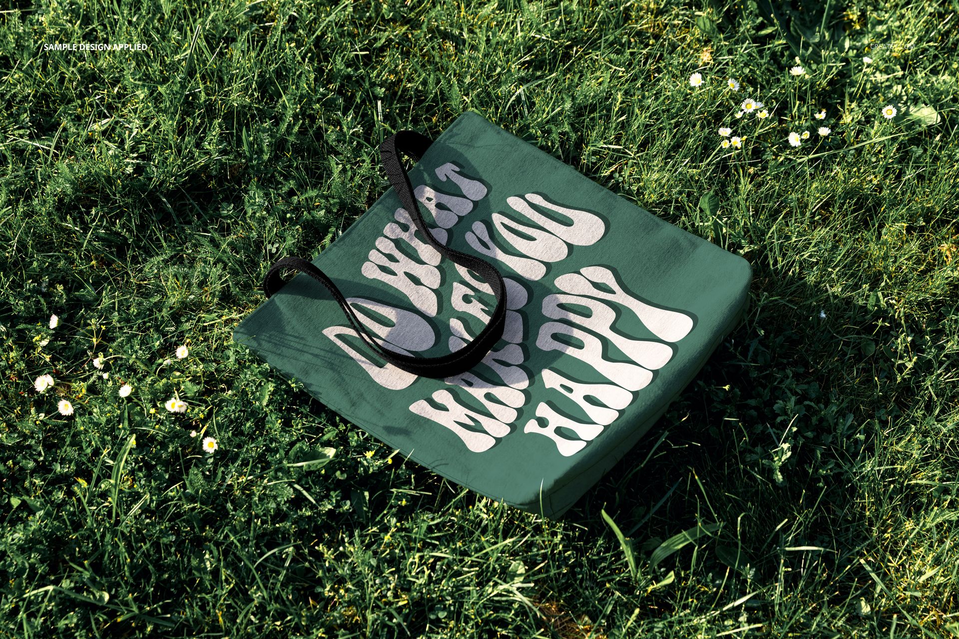 PSD mockup of a woven tote bag with a playful white text pattern, resting among small flowers and blades of grass.