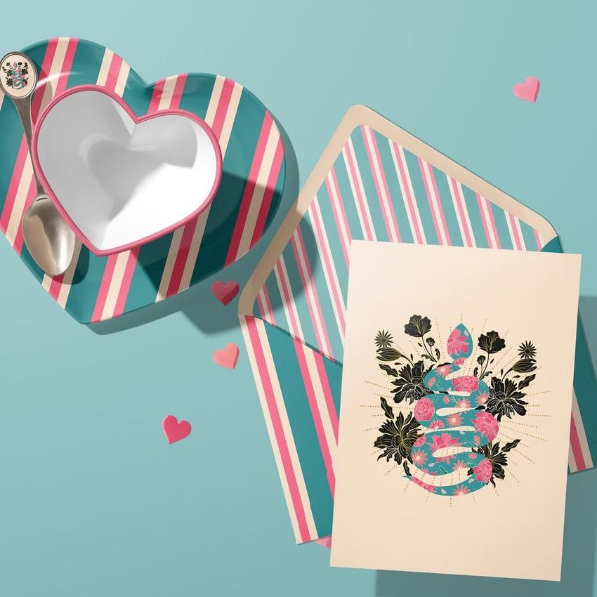 Valentines card mockup featuring a floral snake illustration on cream paper, next to a striped heart-shaped dish and envelope on a blue background.