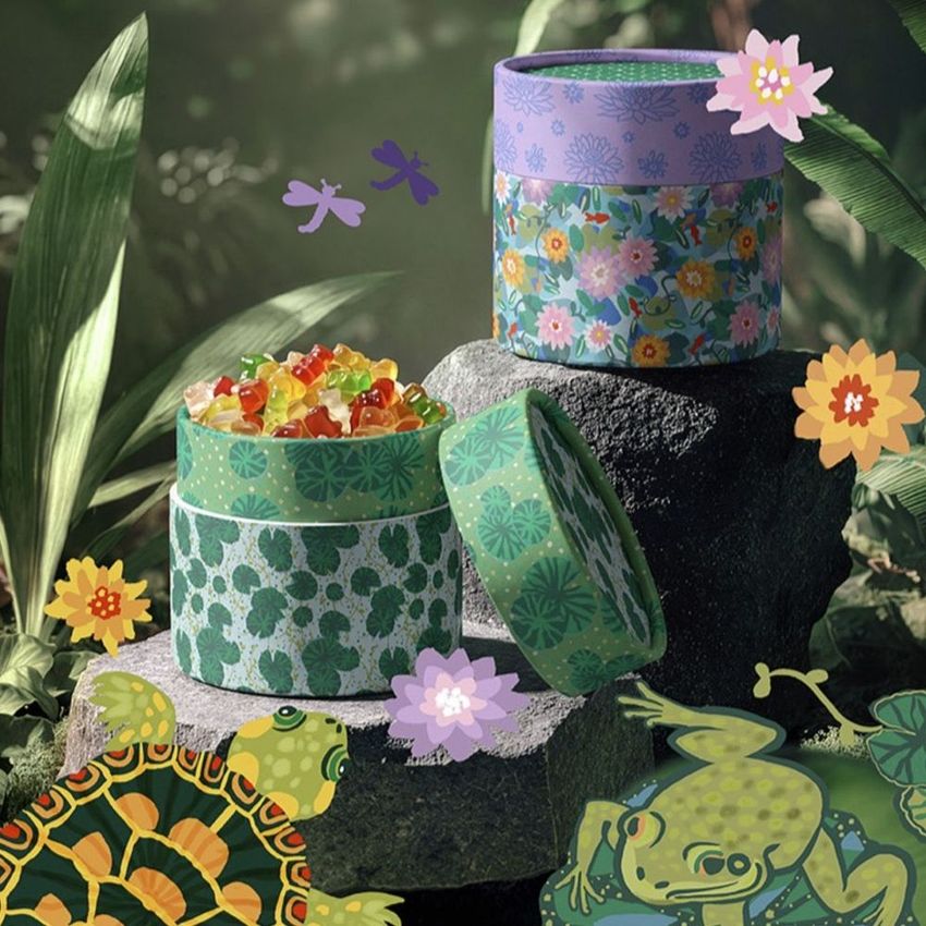 Paper tube packaging mockup featuring colorful floral and geometric patterns, displayed on rocks in a lush jungle setting with gummy candies.