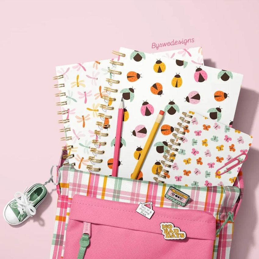 School backpack mockup with spiral notebooks featuring ladybug and dragonfly patterns, pastel pencils, and a plaid bag on a pink background.