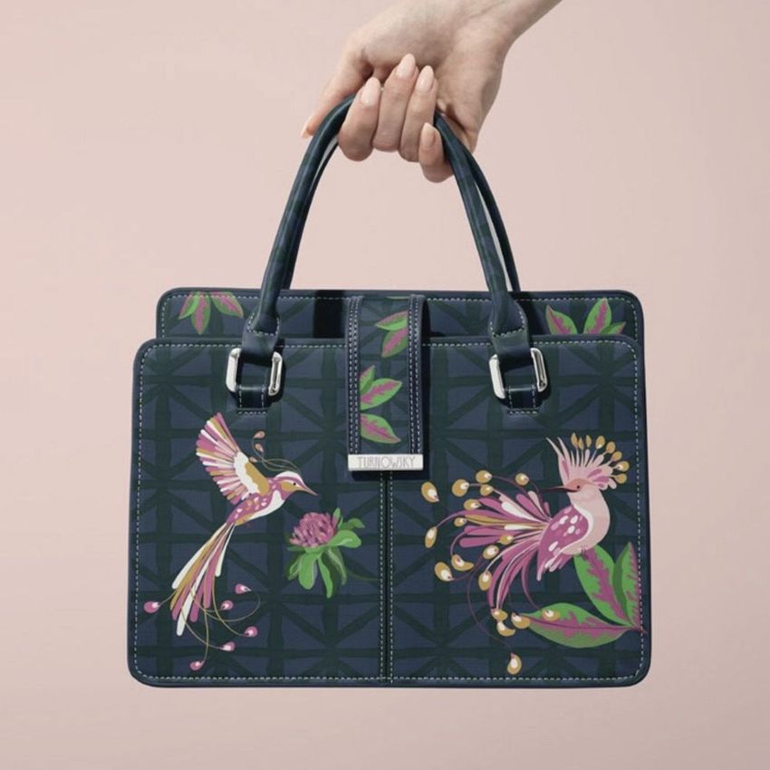 Hand holding a leather handbag mockup with a dark blue base, decorated with colorful bird and floral illustrations on both sides.