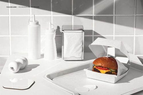 Burger with cheese and vegetables in a white box on a tray, surrounded by condiment bottles and napkin dispenser, mockup scene.