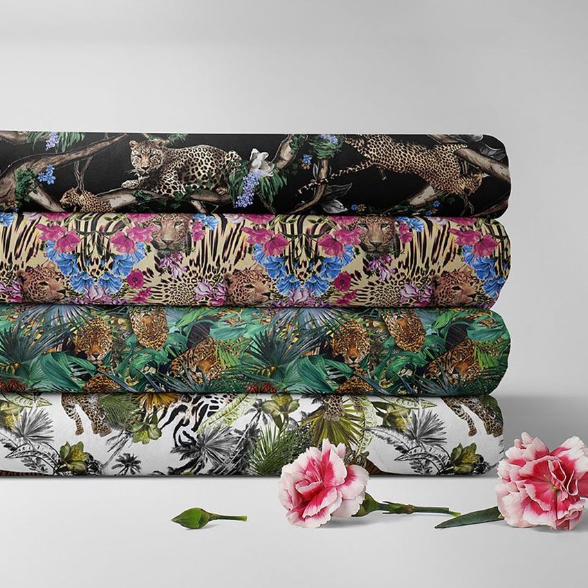 Folded fabrics mockup showing four stacked textiles with jungle animal and floral patterns, set on a light surface with pink flowers.