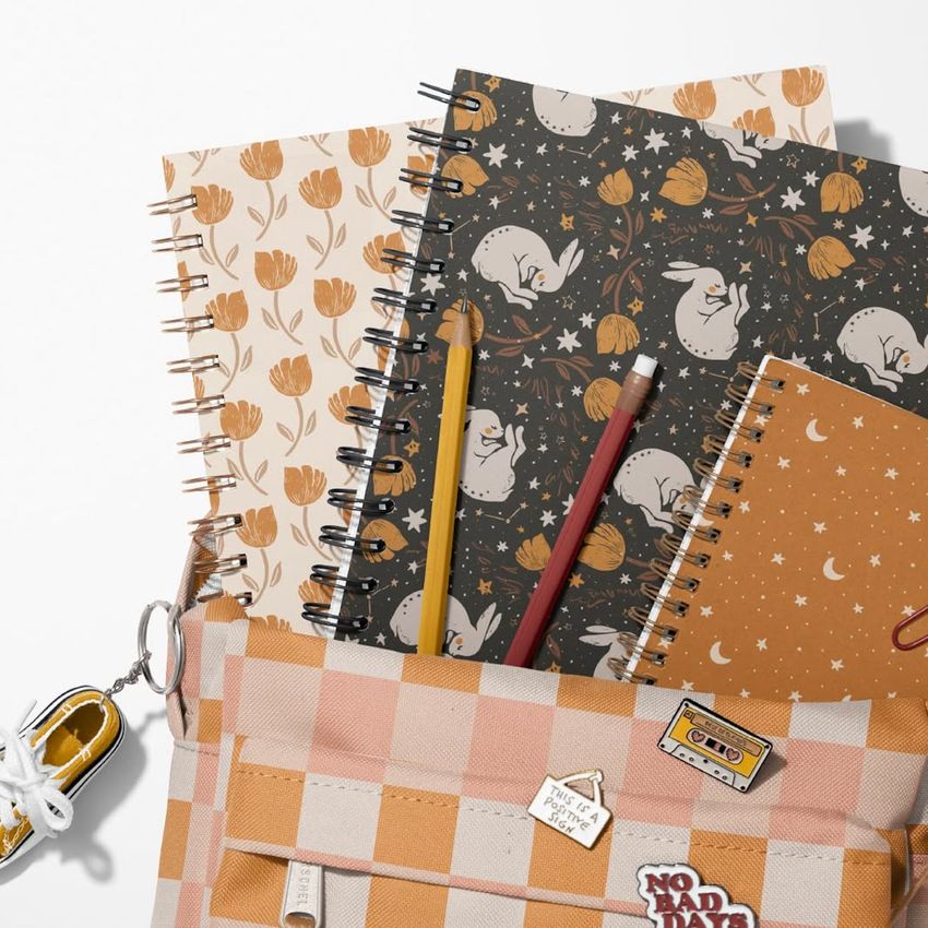 School backpack mockup with spiral notebooks featuring floral, rabbit, and celestial patterns, viewed from above on a white surface.