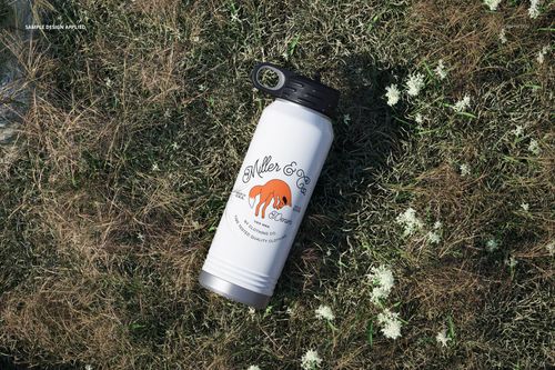 Mockup of a white full color water bottle with black cap, shown from above on grass with scattered white blossoms.