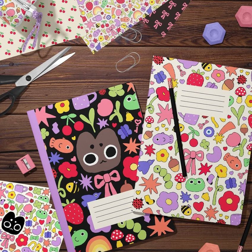 Stationery mockup featuring two A4 notebooks with colorful cartoon covers, surrounded by scissors, paper clips, and patterned paper on a wooden desk.
