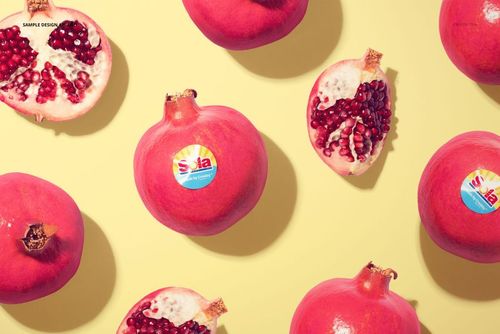Round fruit stickers displayed on whole and halved pomegranates, arranged on a light yellow surface in a mockup scene.