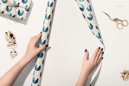 Hands rolling out white gift wrapping paper with blue abstract pattern, surrounded by wrapped gifts and gold scissors.