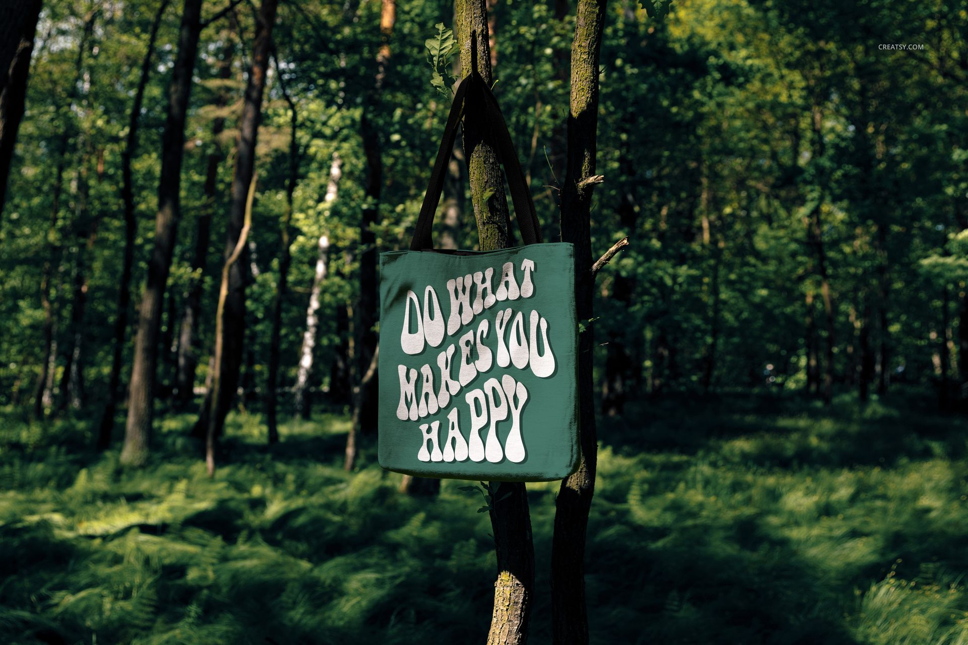 Woven tote bag mockup featuring a green bag with white lettering, displayed outdoors among tall trees and grass.