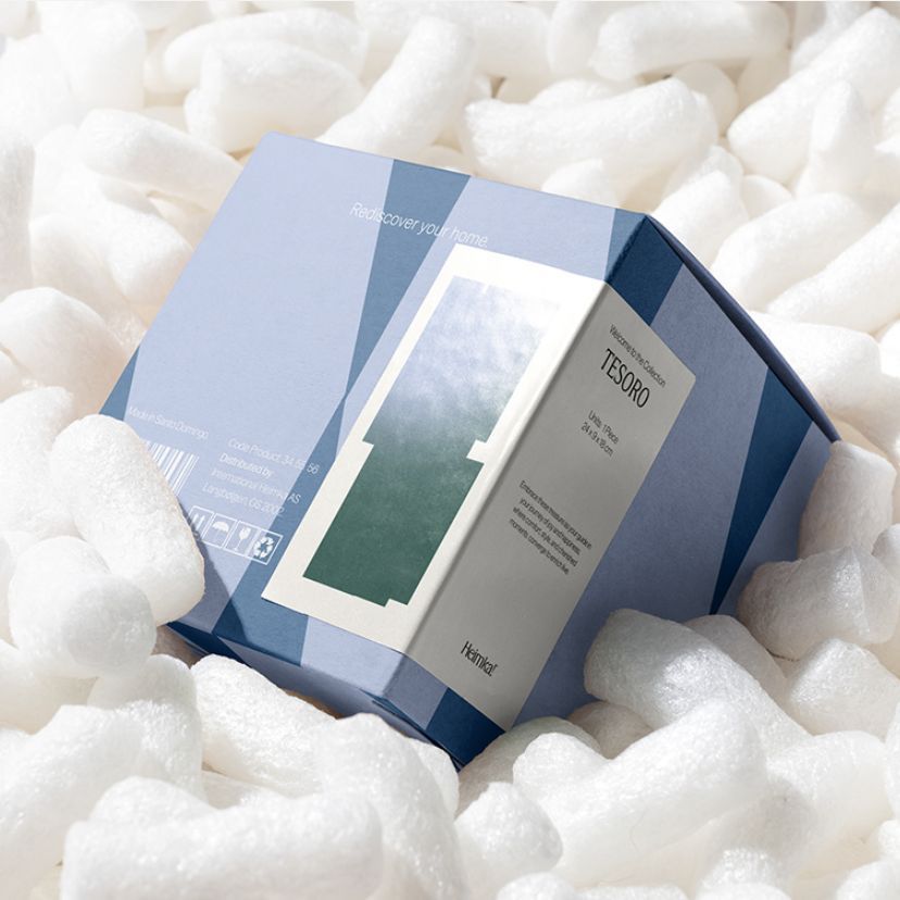 Square box packaging mockup resting on white packing peanuts, with geometric blue design and a visible label on the front side.