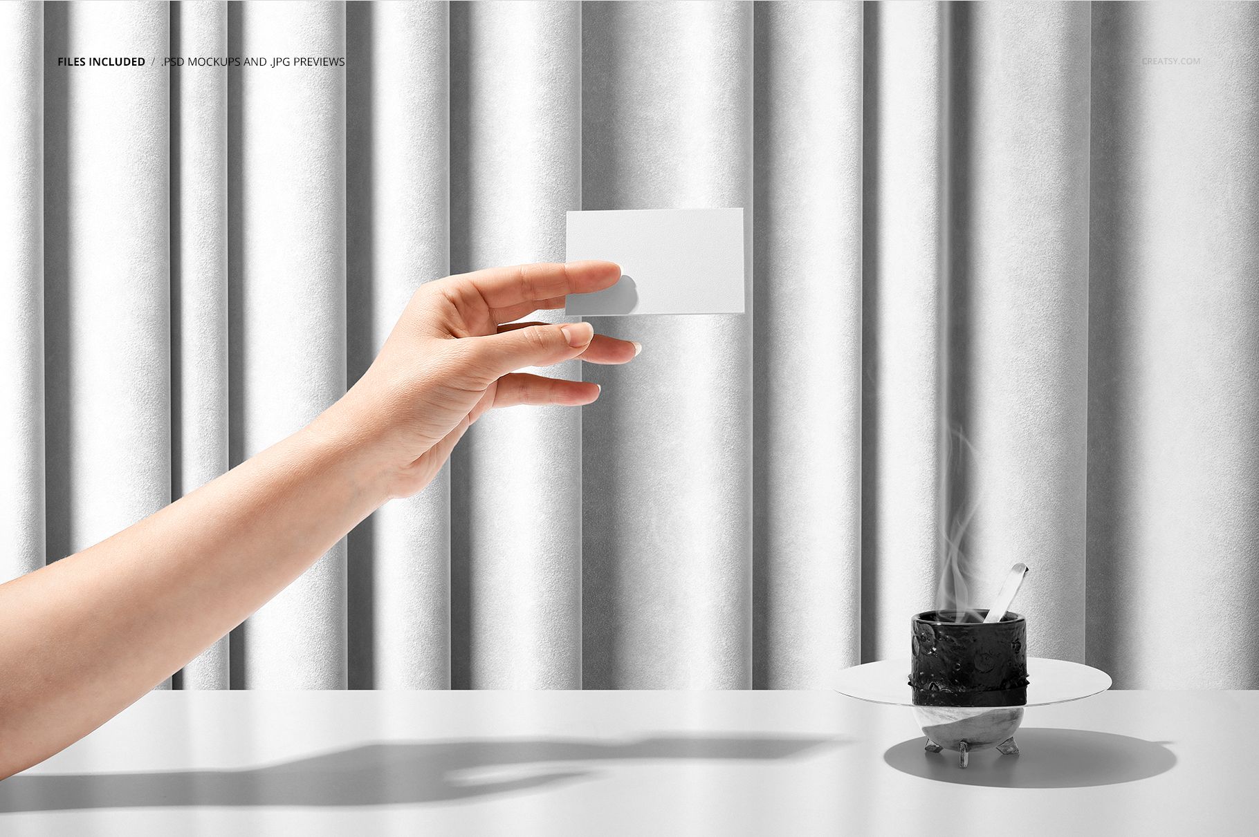 Luxury fabric PSD mockup featuring vertical silver textile folds, a hand with a card, and a steaming cup on a table.