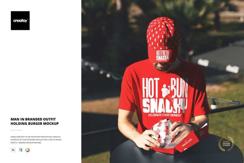 Front view of a man in a red graphic t-shirt and matching cap, holding a burger box mockup in a sunny outdoor setting.