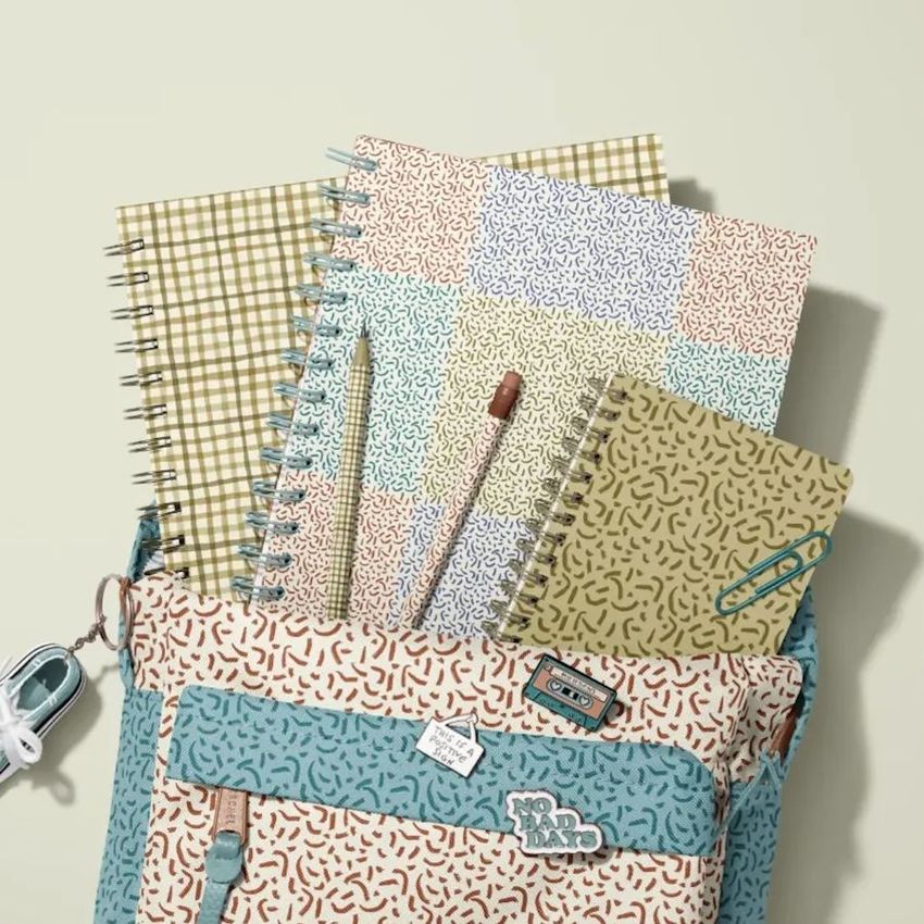 School backpack mockup with spiral notebooks featuring abstract and plaid patterns, a pencil, and a keychain on a light surface.