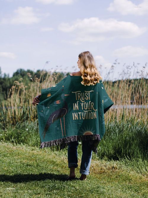 Outdoor scene featuring a woven poncho mockup with illustrated pattern and text, displayed against a natural lakeside backdrop.