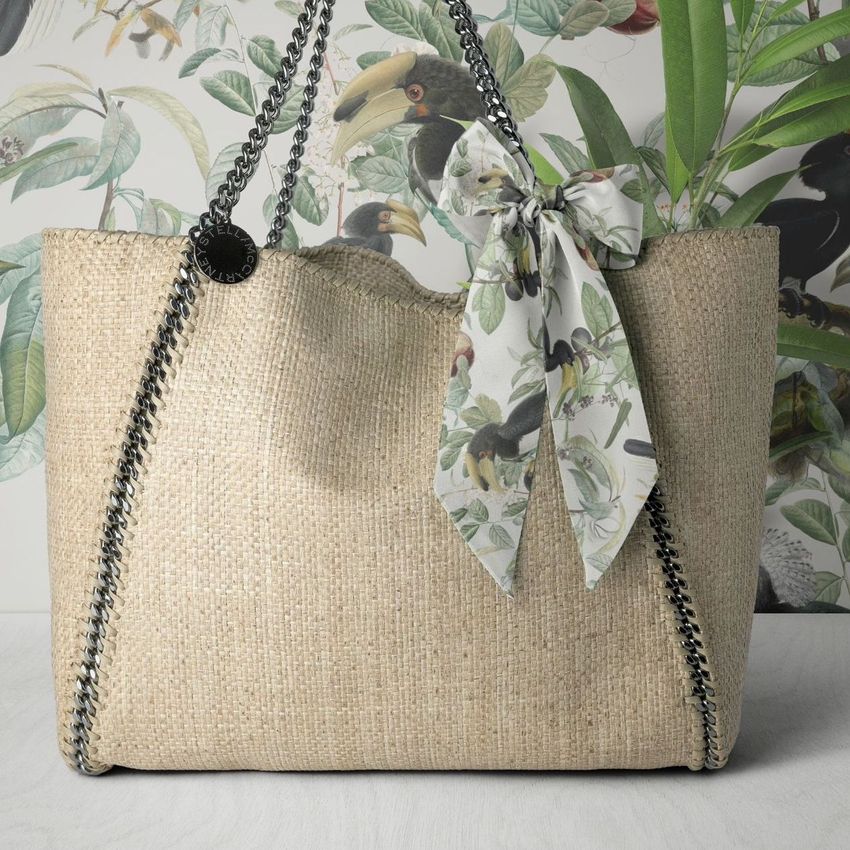Twilly scarf mockup featuring a light patterned scarf tied to the handle of a woven beige bag, set against a tropical wallpaper.