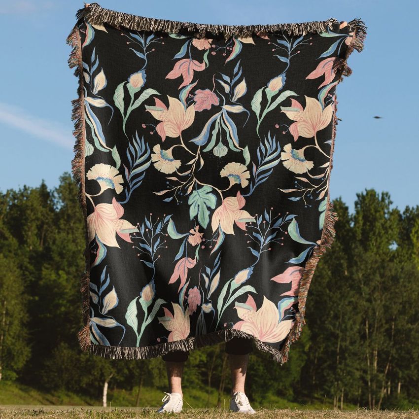 Person holding up a woven throw blanket mockup with a floral pattern in pastel colors on a black background outdoors.