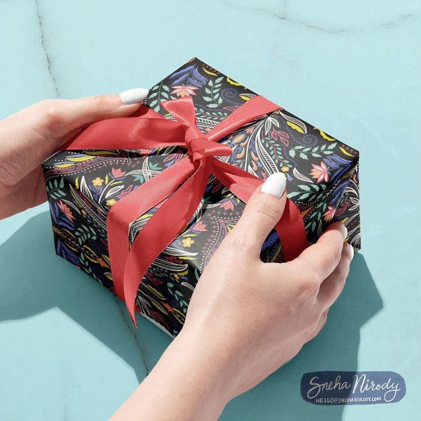 Gift box mockup featuring dark floral wrapping paper with colorful folk-inspired patterns and a red ribbon, held by two hands on a light blue surface.