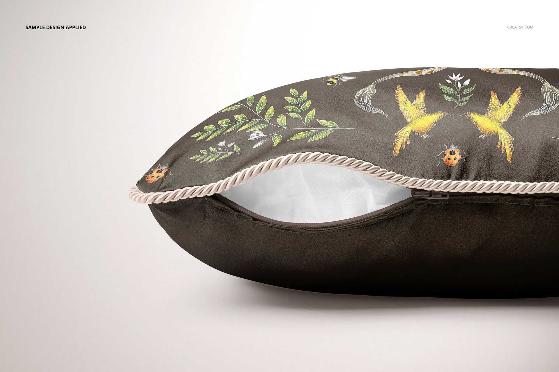 Close-up of a silk cushion mockup with braid detail, decorated with leaves, birds, and ladybugs on a dark fabric.