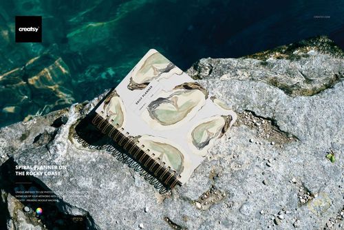 Stationery mockup showing a spiral-bound planner with artistic cover placed on textured rocks by the water.