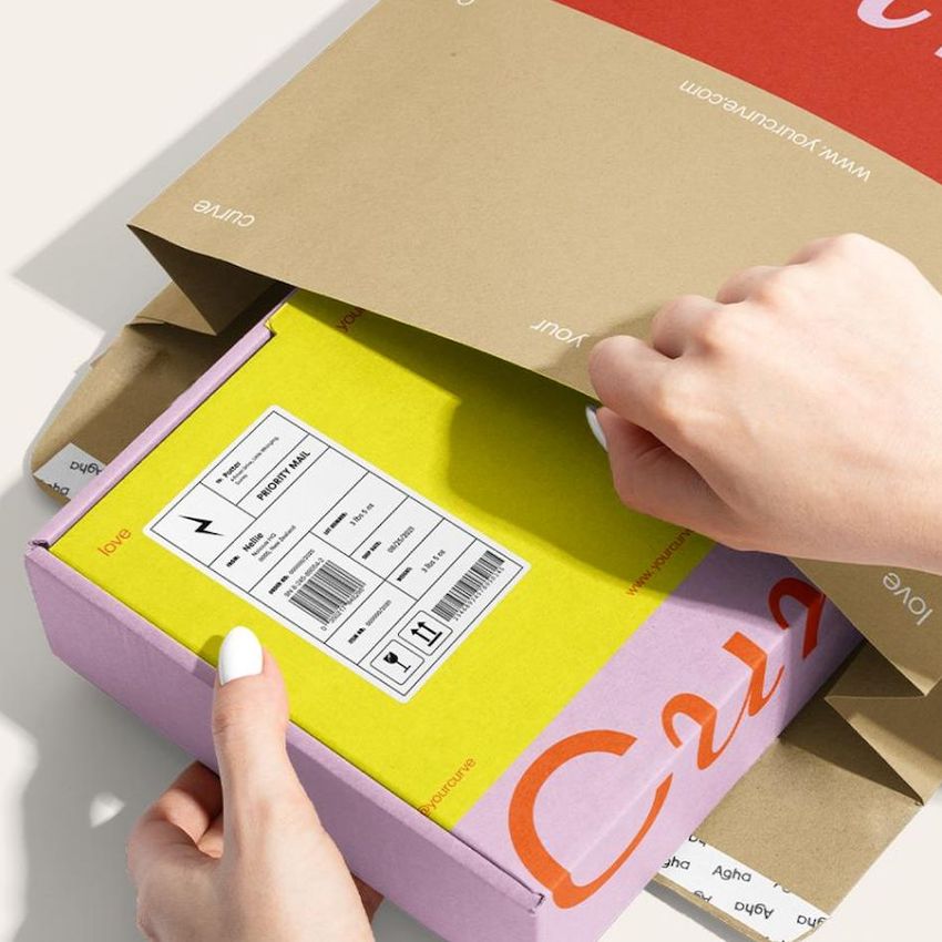 Hands opening a brown kraft mailer with a red and white design, revealing a yellow and purple box with a shipping label.