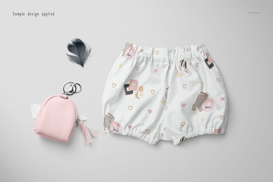 Baby harem shorts mockup featuring a soft white fabric with cartoon bears and hearts, shown beside a feather and small bag.