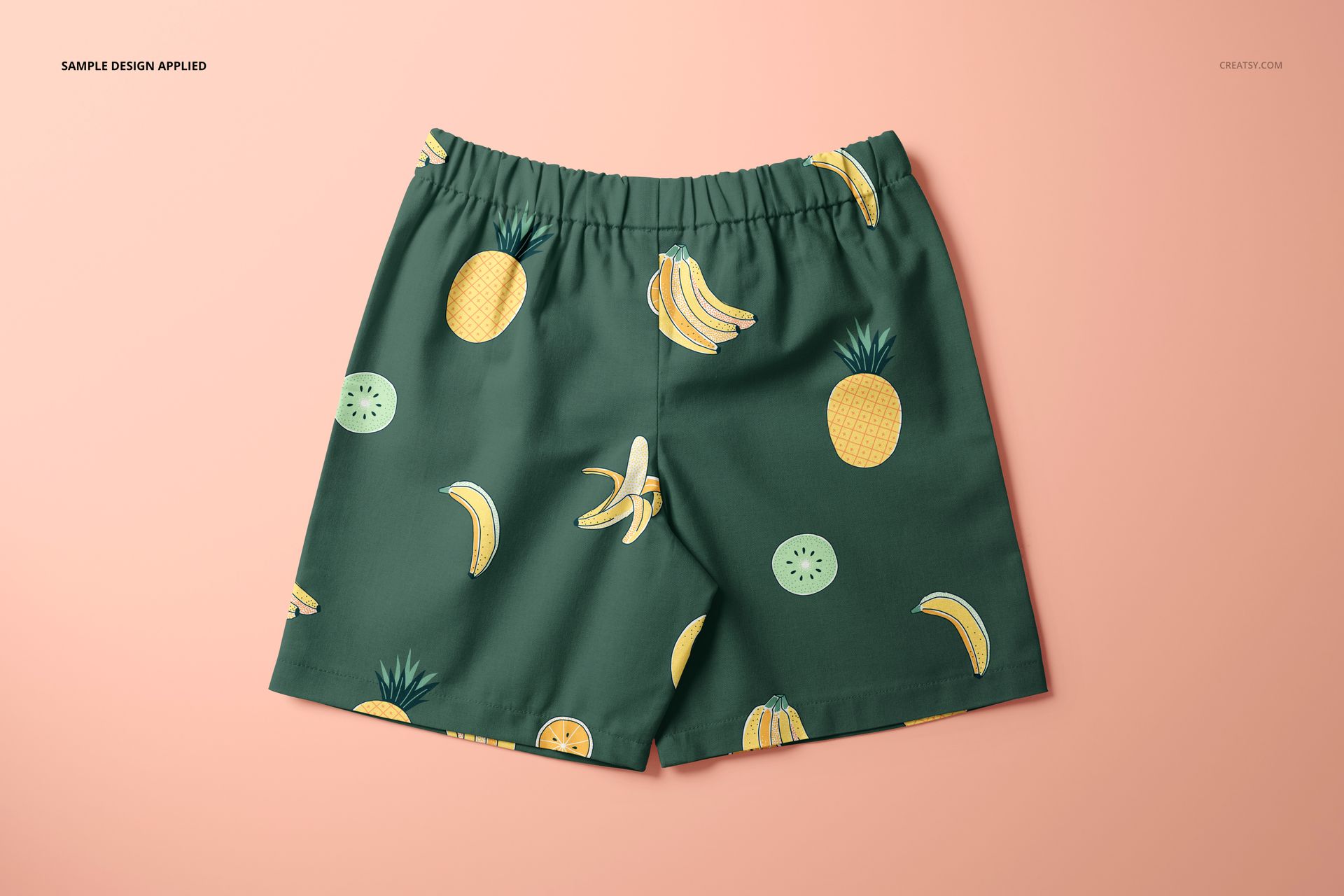 Kids shorts mockup featuring a green fabric with playful fruit illustrations, shown from a top front view on a peach background.