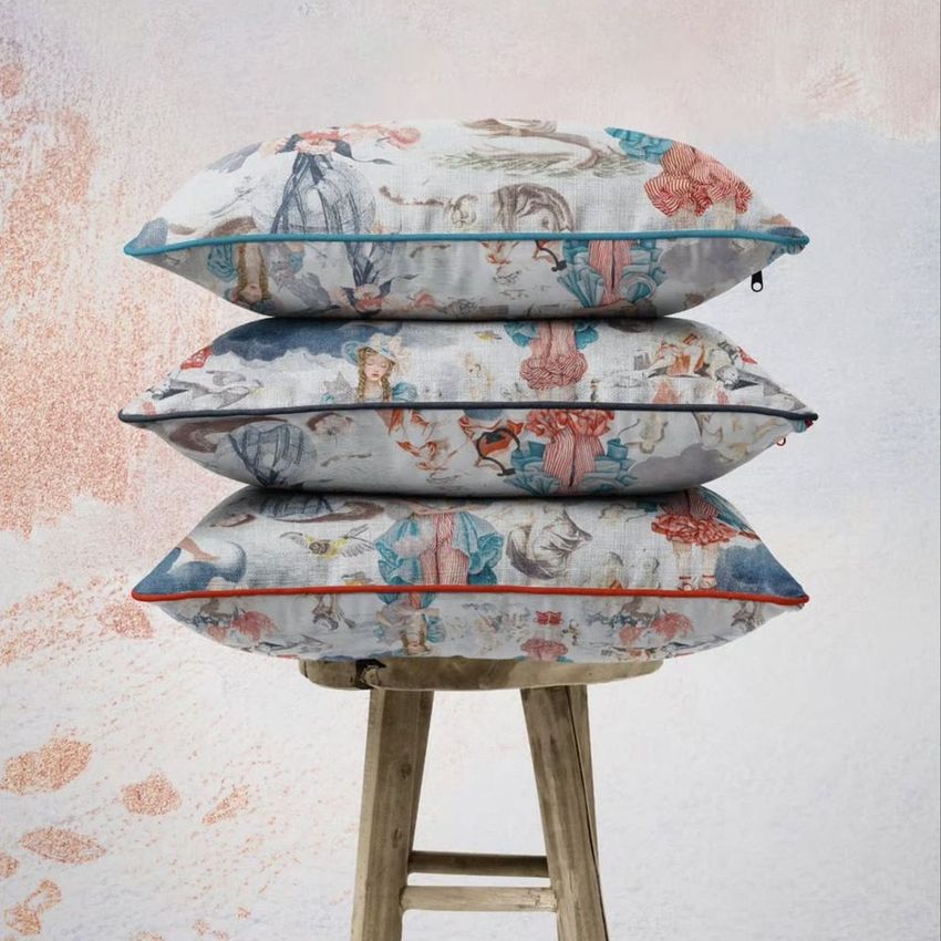 Stack of three reversible piped cushions mockup with intricate rococo-style illustrations, placed on a rustic wooden stool.