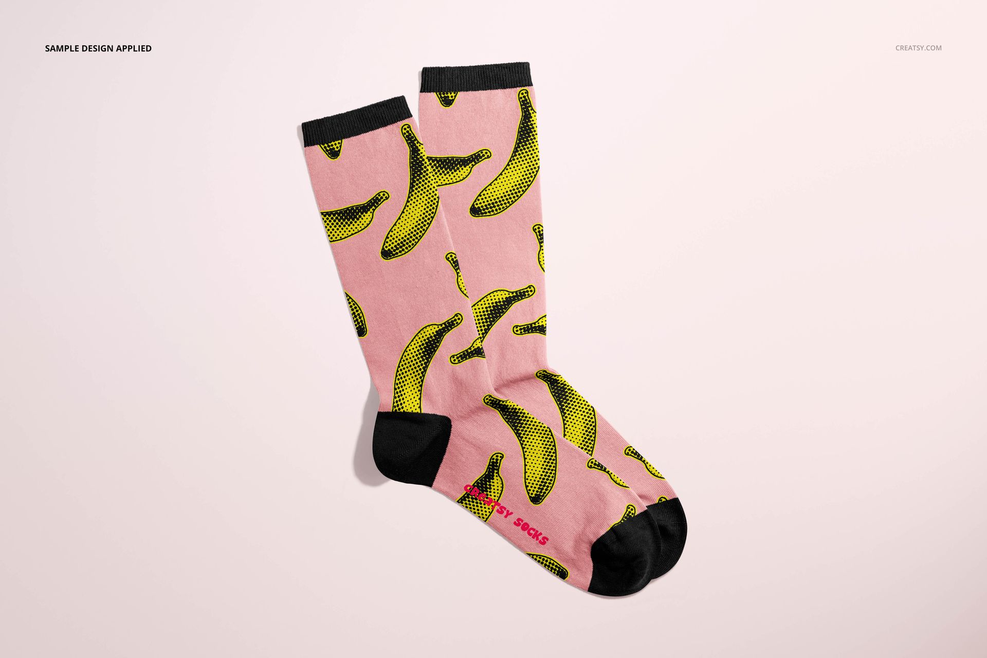 Front view of two socks in a mockup scene, pink fabric with black accents and yellow banana illustrations.