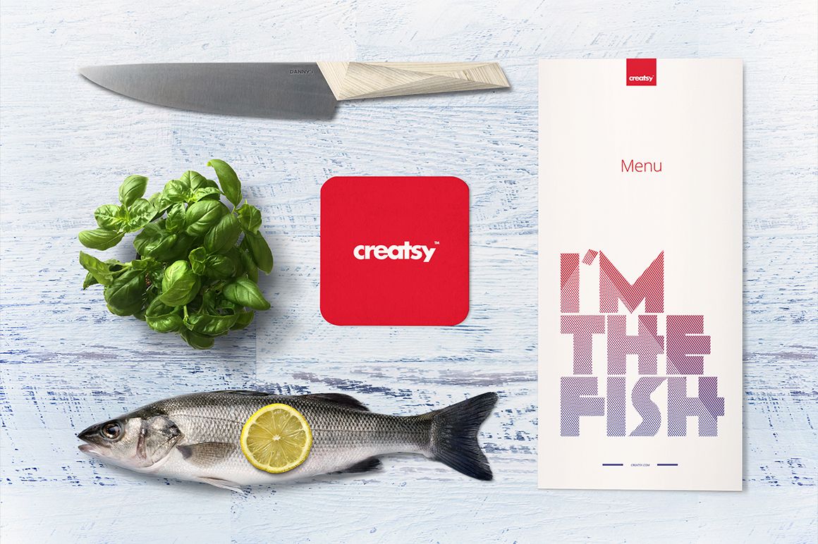 Seafood restaurant mockup featuring a fish with lemon slice, menu, knife, basil leaves, and red coaster on a white wooden surface.