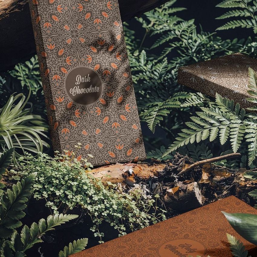 Chocolate box mockup featuring ornate brown packaging with orange accents, placed among lush green forest foliage and logs.