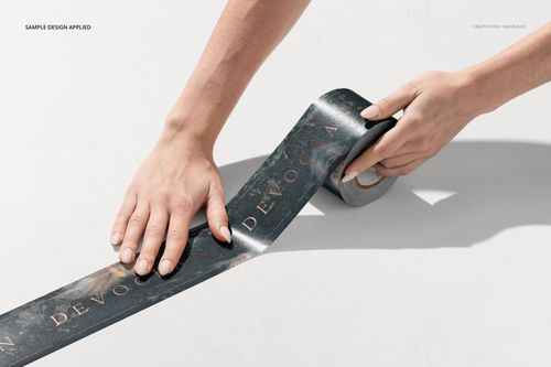 Hands applying a dark patterned adhesive tape with gold text on a white surface, viewed from above.